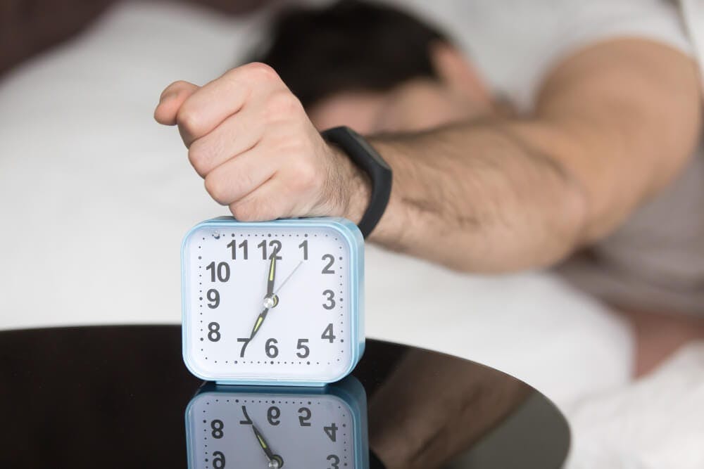 Angry sleepy young guy turning off noisy annoying alarm clock because he is oversleeping 